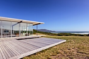 A modern house with a wooden deck and large glass windows overlooking the ocean. - Cloudy Bay Beach House (South Bruny)