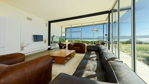 A modern living room with a large sectional sofa, a coffee table, and a TV on a white wall. There is a large window offering a view of the outdoors.