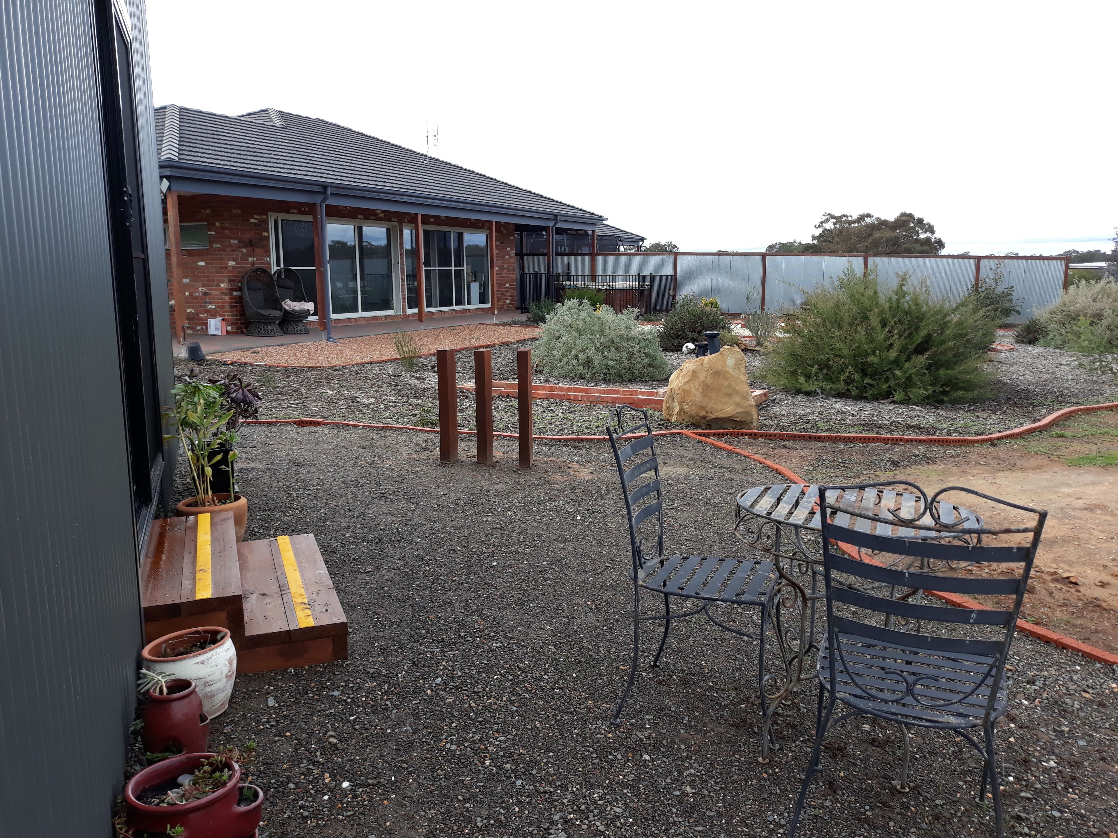 A patio area with a table and chairs, a brick building with large windows, and a garden with various plants and a hose.