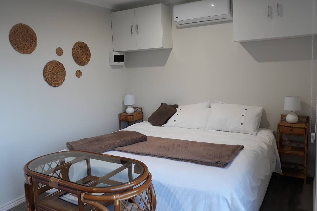 Un dormitorio con una cama, dos mesitas de noche, una silla de madera pequeña y un aire acondicionado de pared.