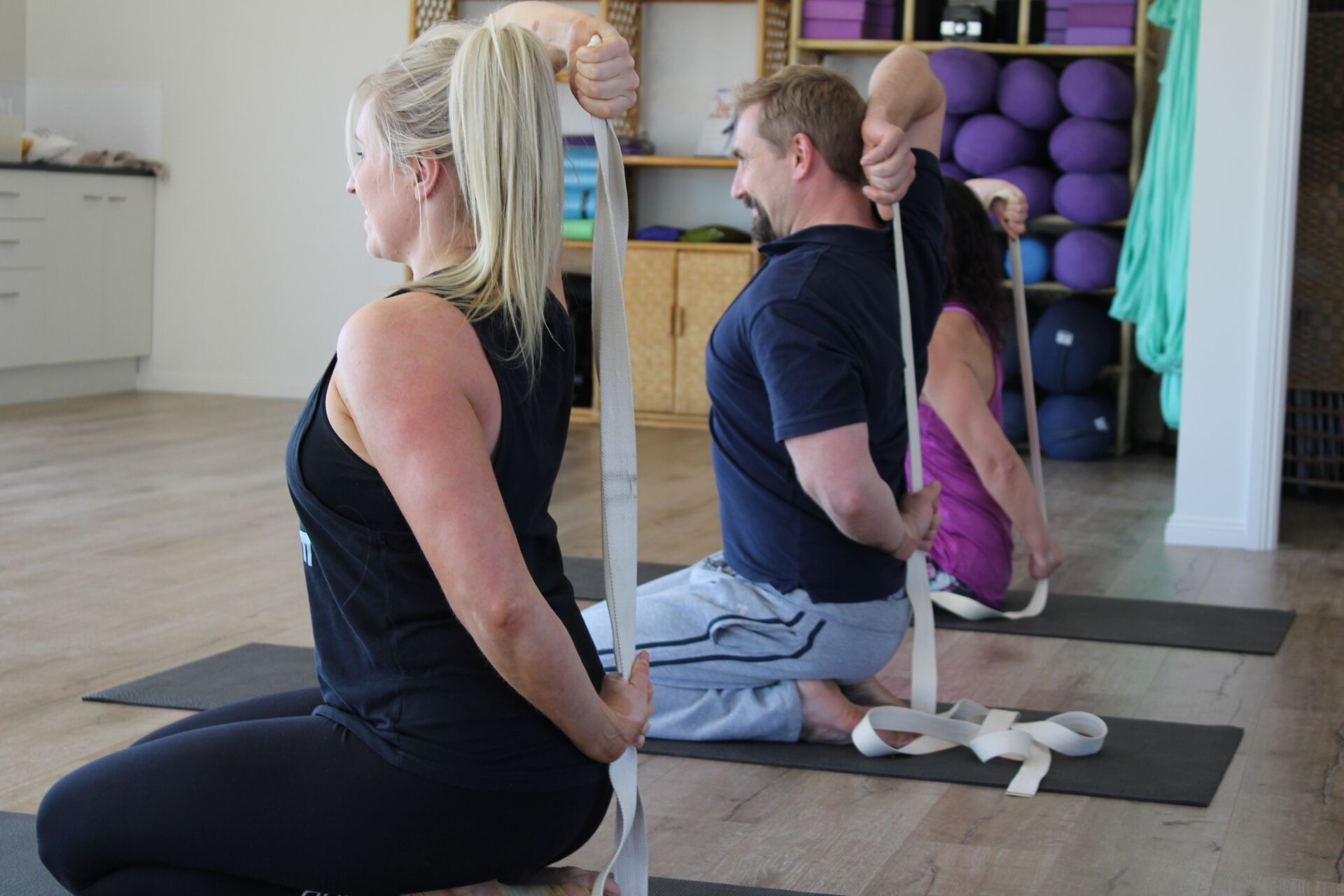 A yoga class in a room with wooden flooring, exercise mats, and a shelf with yoga balls.