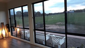 Ein Zimmer mit großen Fenstern mit Blick auf ein saftiges Grasfeld, Holzboden und ein weißer Kunststoffstuhl.