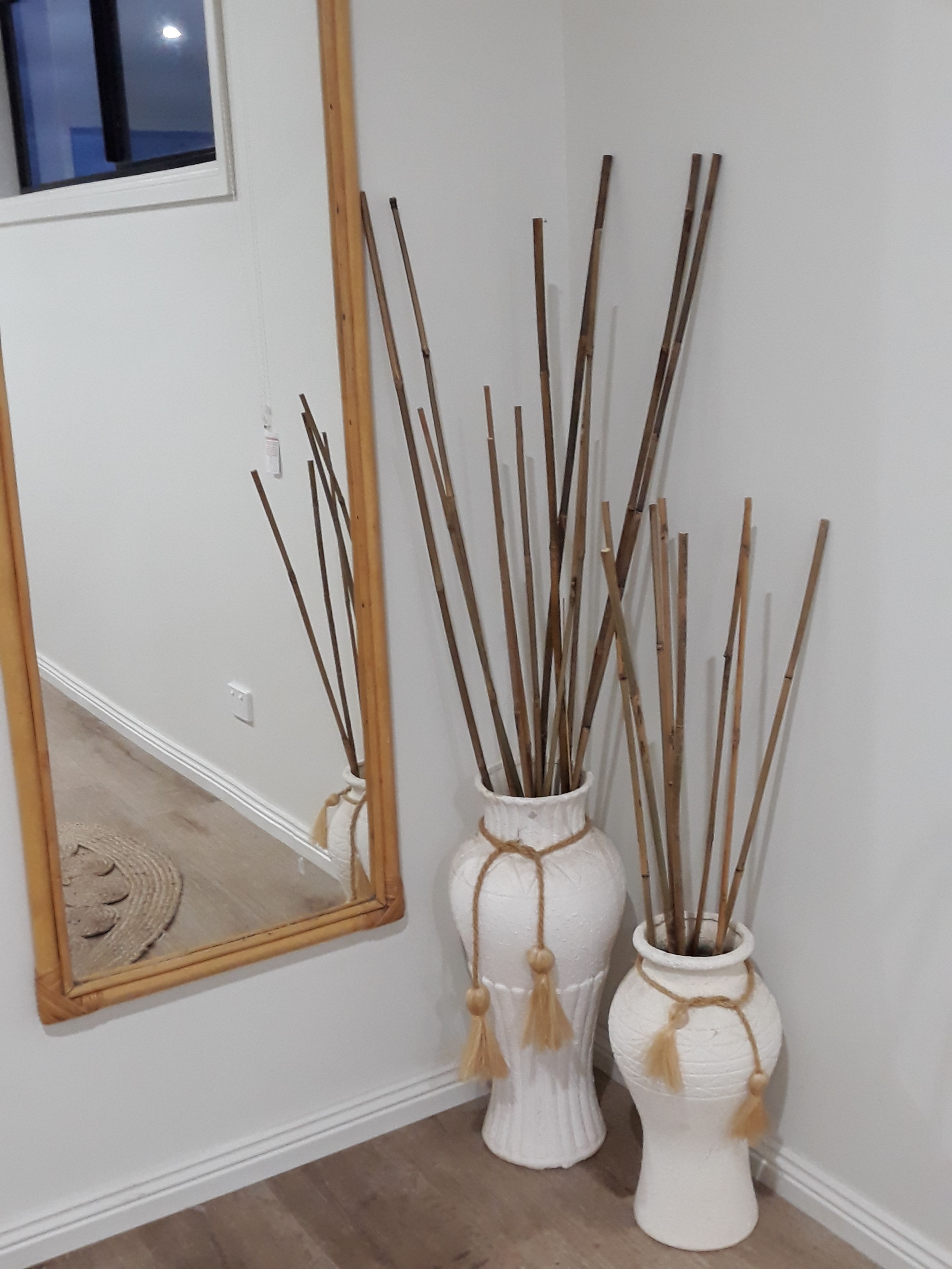 A room with a large mirror reflecting a window, two white vases with wooden sticks and tassels, and a wooden floor.