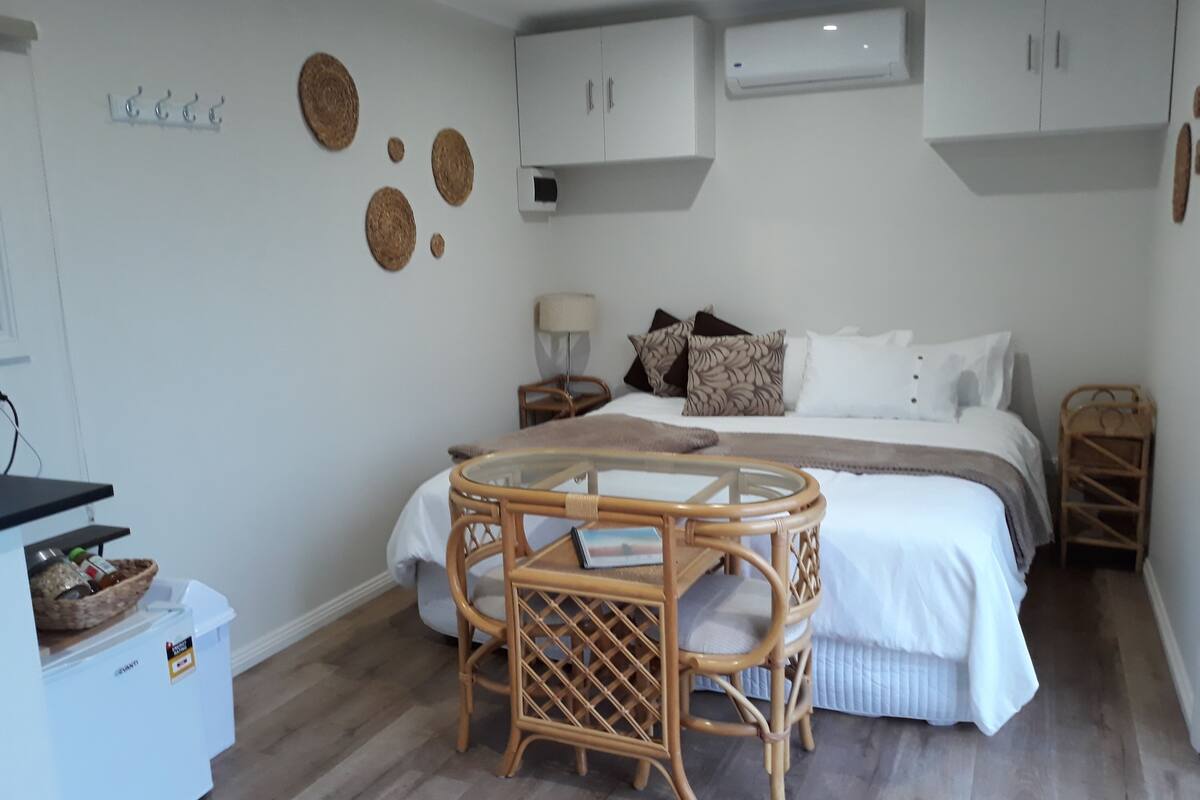Un dormitorio con cama, mesitas de noche, una mesa de vidrio, sillas de mimbre y un armario blanco.