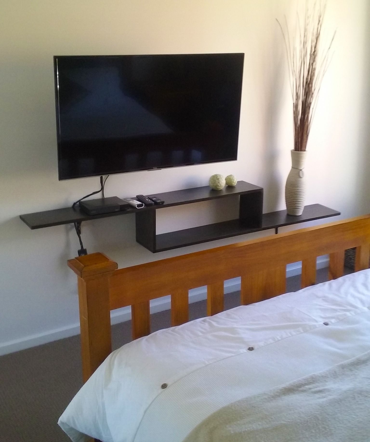 Ein Schlafzimmer mit einem Bett, einem an einer Ablage montierten Fernseher und einer Vase mit Trockenpflanzen.