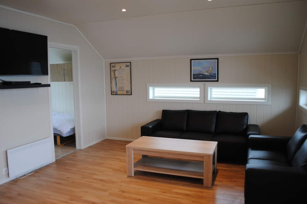 A modern living room with a wooden floor, black leather sofa, a wooden coffee table, a flat-screen TV mounted on the wall, and a framed picture above the sofa.