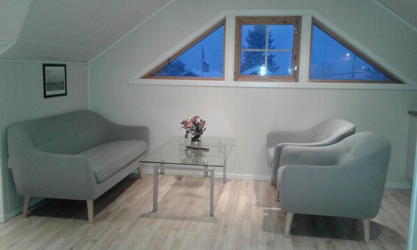 A modern living room with a glass coffee table, grey sofa, and armchairs. A wooden window with a view of trees is above the table. - Furoy Hotel Apartments and Boating (Sorreisa)