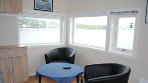 A room with a wooden cabinet, a table with a blue tablecloth, two black chairs, and large windows overlooking a body of water. - Furoy Hotel Apartments and Boating (Sorreisa)