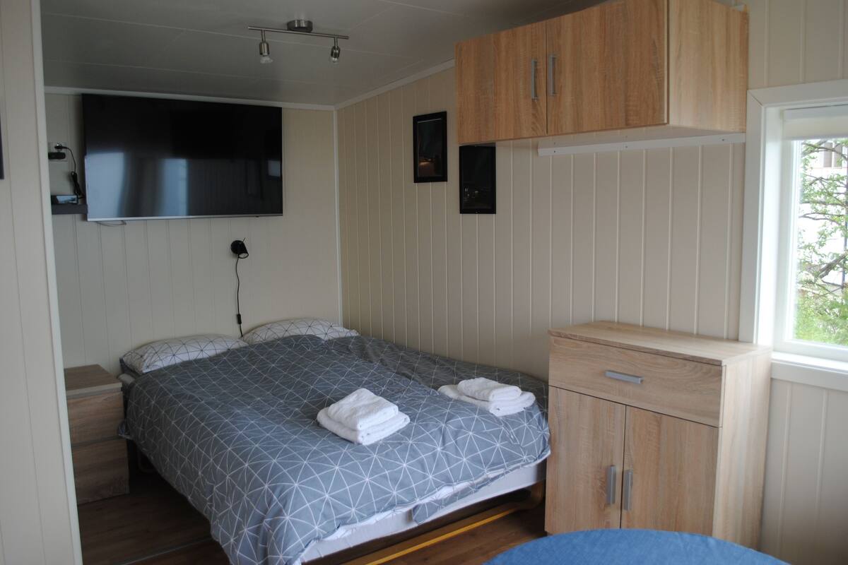 A bedroom with a bed, a TV mounted on the wall, a wooden cabinet, and a window.