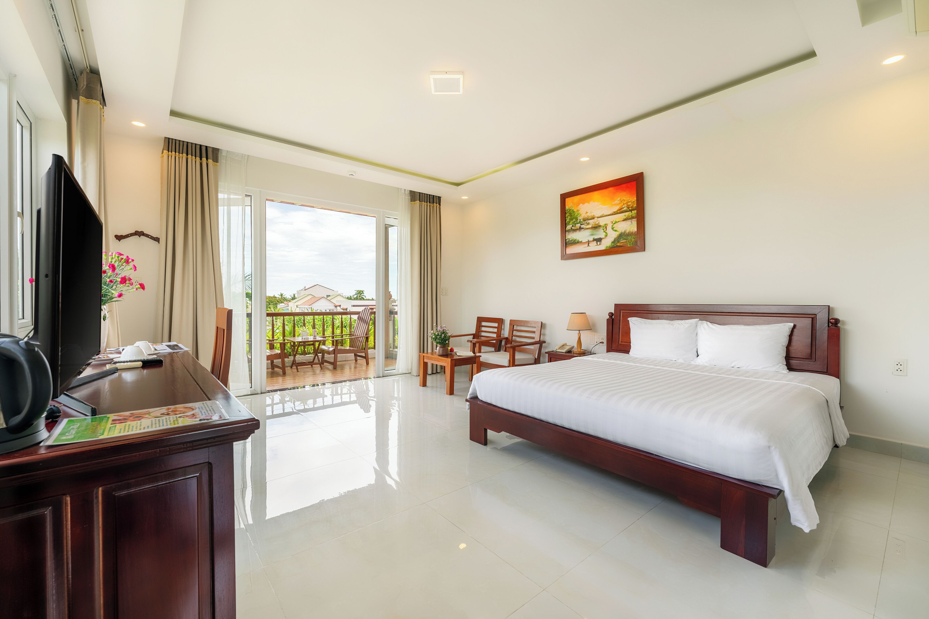 A hotel room with a large bed, a desk with a television, and a balcony with outdoor seating.