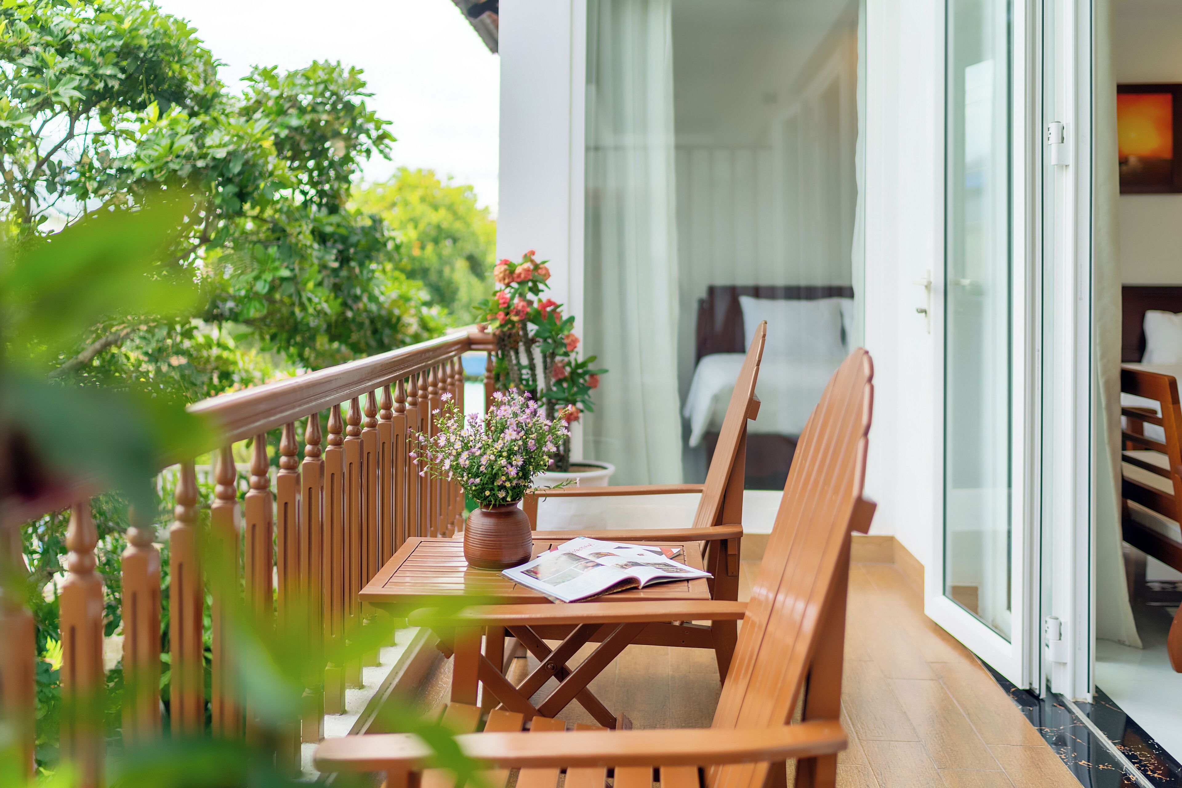 A balcony with wooden furniture, a potted plant, and a view of a bedroom with a bed and curtains.