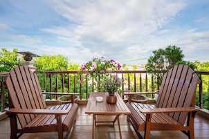 Una terraza de madera con dos sillas y una mesa, rodeada por una barandilla con una planta en maceta y flores.