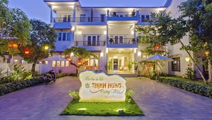 Un hotel de diverses plantes amb una entrada ben il·luminada, un cartell de 'THINH HUNG Palmy Villa Bar & Restaurant' i un camí pavimentat amb vegetació.