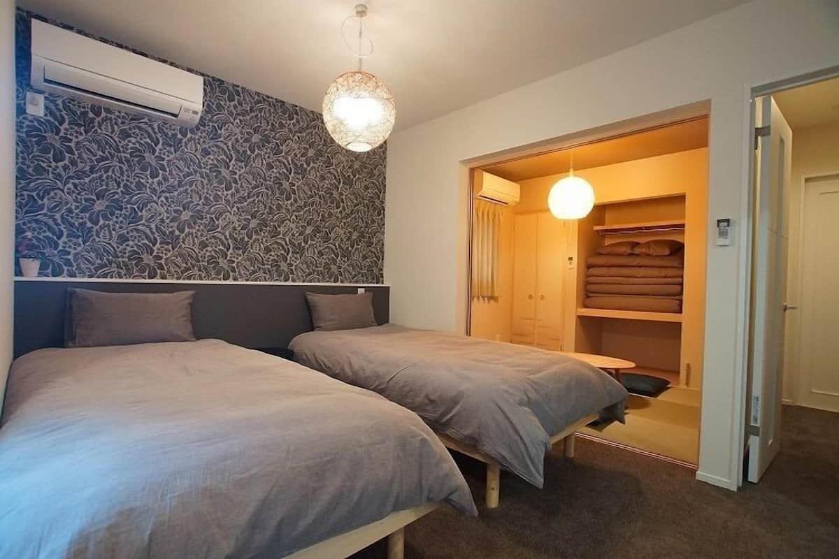 A room with two beds, a patterned wall, an air conditioning unit, a hanging light fixture, and a closet with folded blankets.