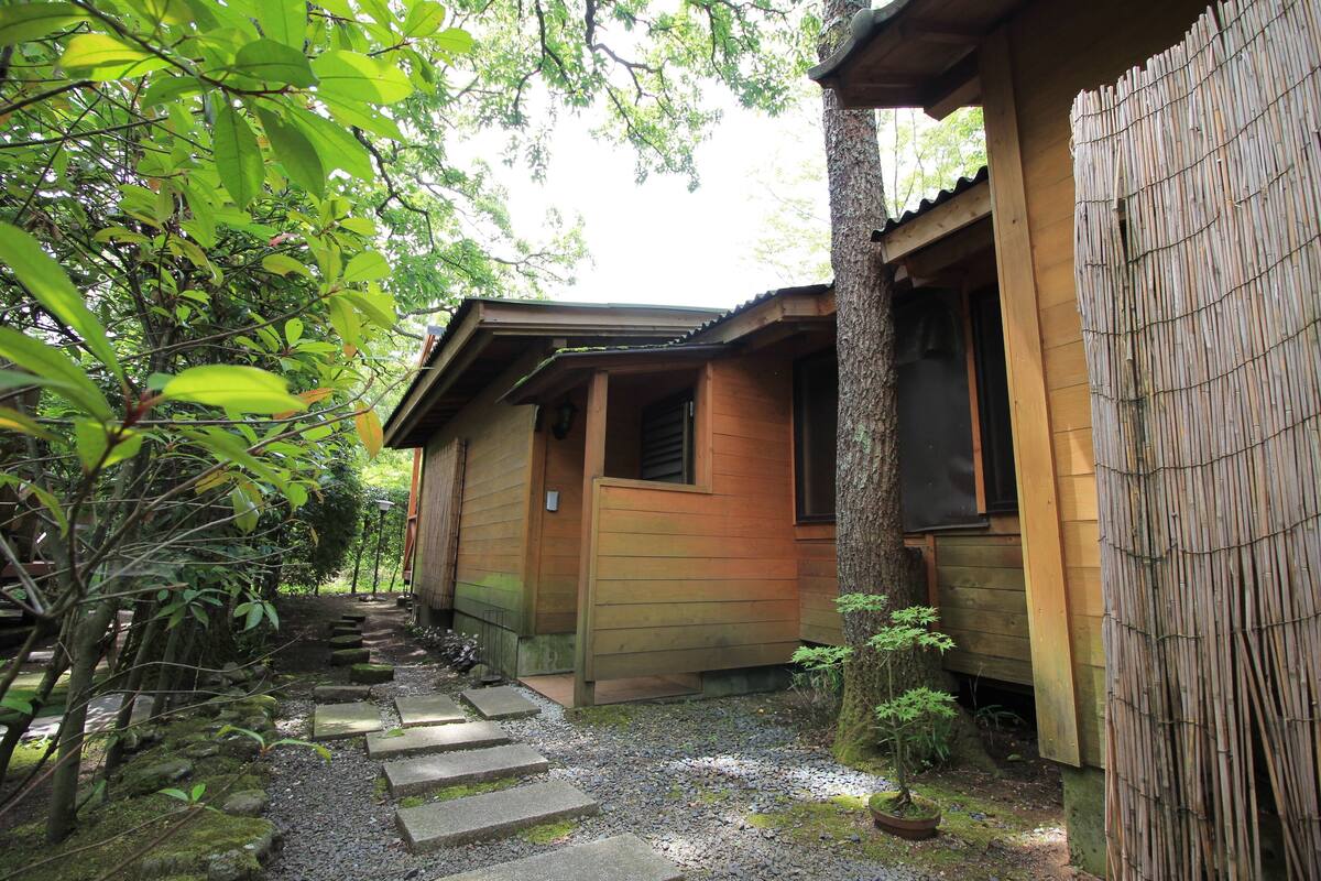Un bâtiment traditionnel en bois, doté d’une porte et de fenêtres en bois, entouré d’un chemin de pierre et de verdure.