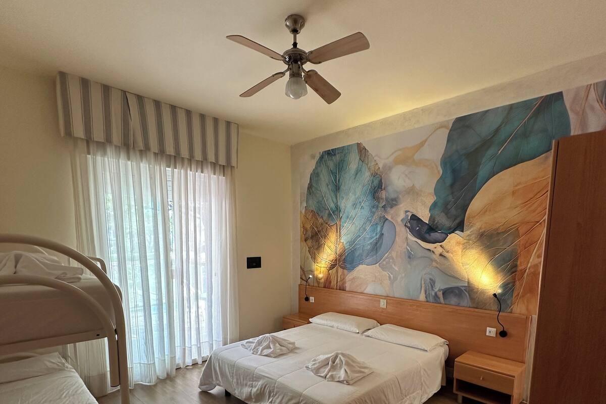 A hotel room with a bed, a ceiling fan, a wooden nightstand, a bedside table, and a large abstract painting on the wall.