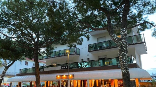 Een meerlaags hotel met een restaurant op de begane grond, terras buiten en een balkon op de bovenste verdieping.