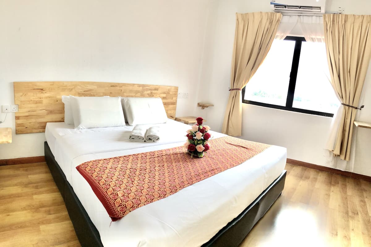 A neatly made bed with white linens, a wooden headboard, and a red patterned bedspread. There is a small wooden nightstand with a phone and a power outlet. A window with beige curtains is visible, and a small flower arrangement is on the bed.