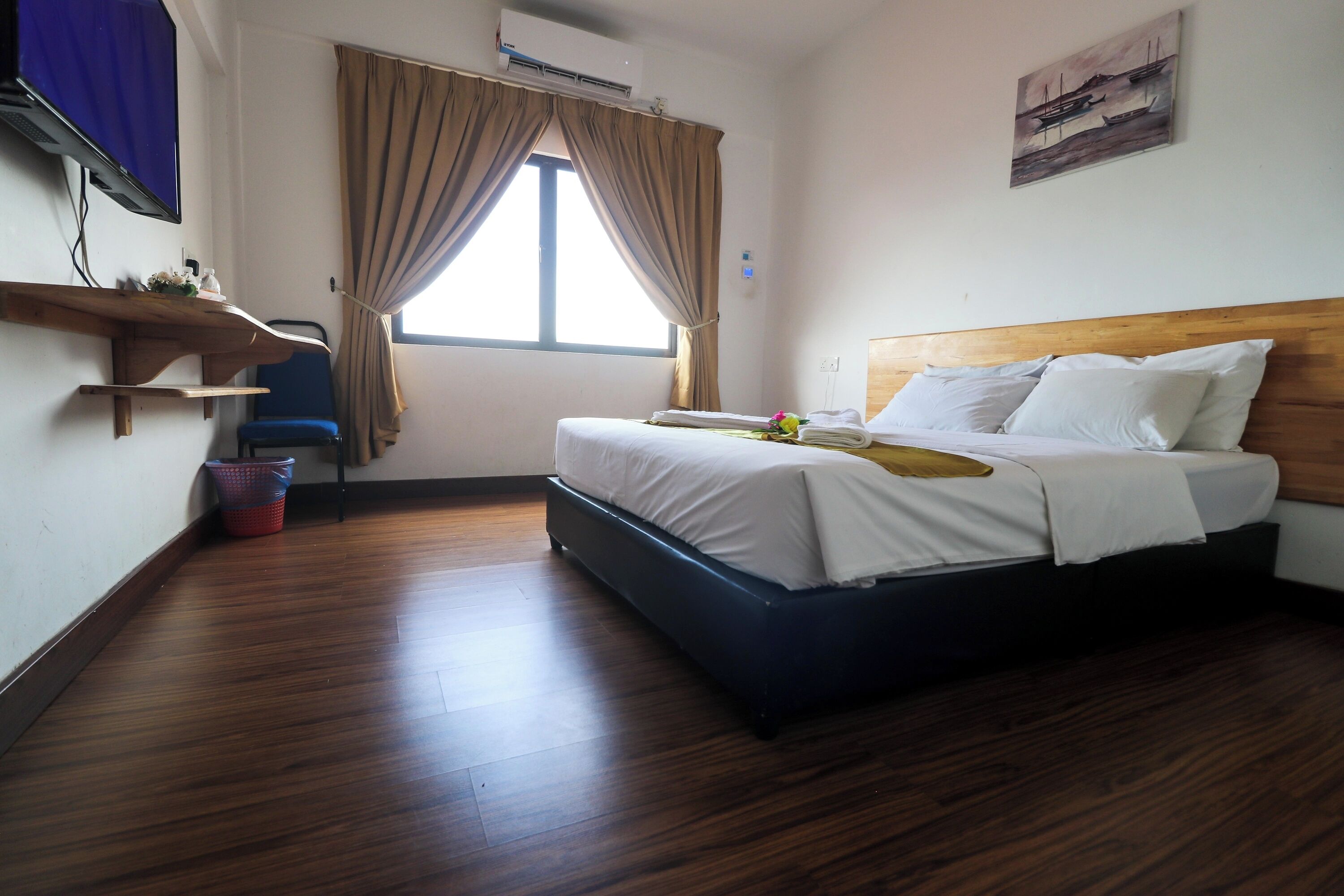 A hotel room with a bed, a TV, a window with curtains, a wall-mounted air conditioner, and a painting.