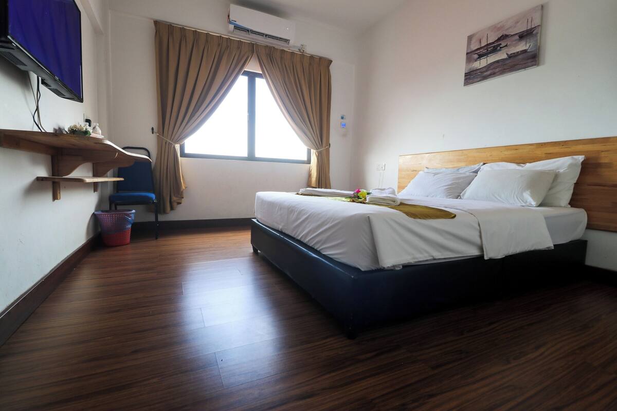 A hotel room with a bed, a TV, a window with curtains, a wall-mounted air conditioner, and a painting.