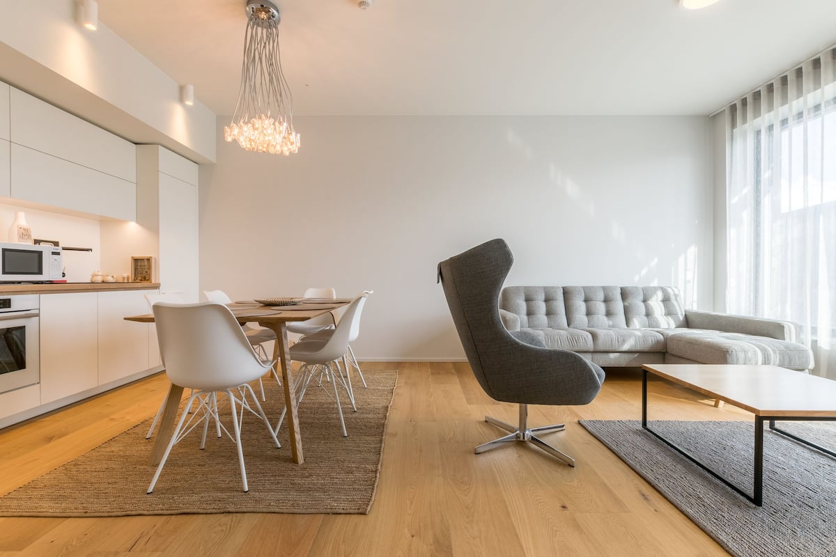 A modern living room with a grey sofa, a dining area with white chairs, and a kitchen with white cabinets.
