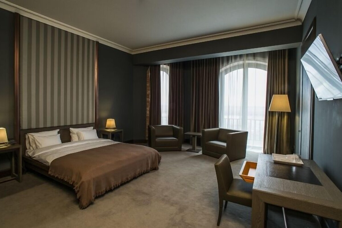 Habitación de hotel con una cama, dos mesitas de noche con lámparas, un escritorio con silla, una pequeña mesa y un ventanal con cortinas.