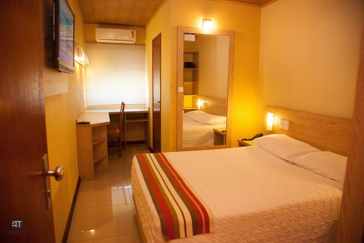 A hotel room with a bed, a desk, a chair, a mirror, and a TV on the wall.