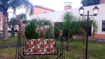 Un jardín con una hamaca, palmeras y un edificio de fachada blanca y tejado rojo.