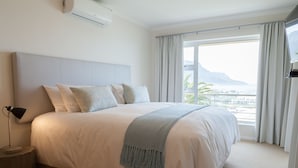 A large bed with a headboard, a bedside lamp, and a view of a balcony with curtains. - Bay Vista Villa (Cape Town)