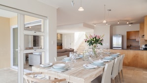 A spacious dining area with a long wooden table set for six, surrounded by wicker chairs. A kitchen is visible in the background with wooden cabinets and a refrigerator. - Bay Vista Villa (Cape Town)