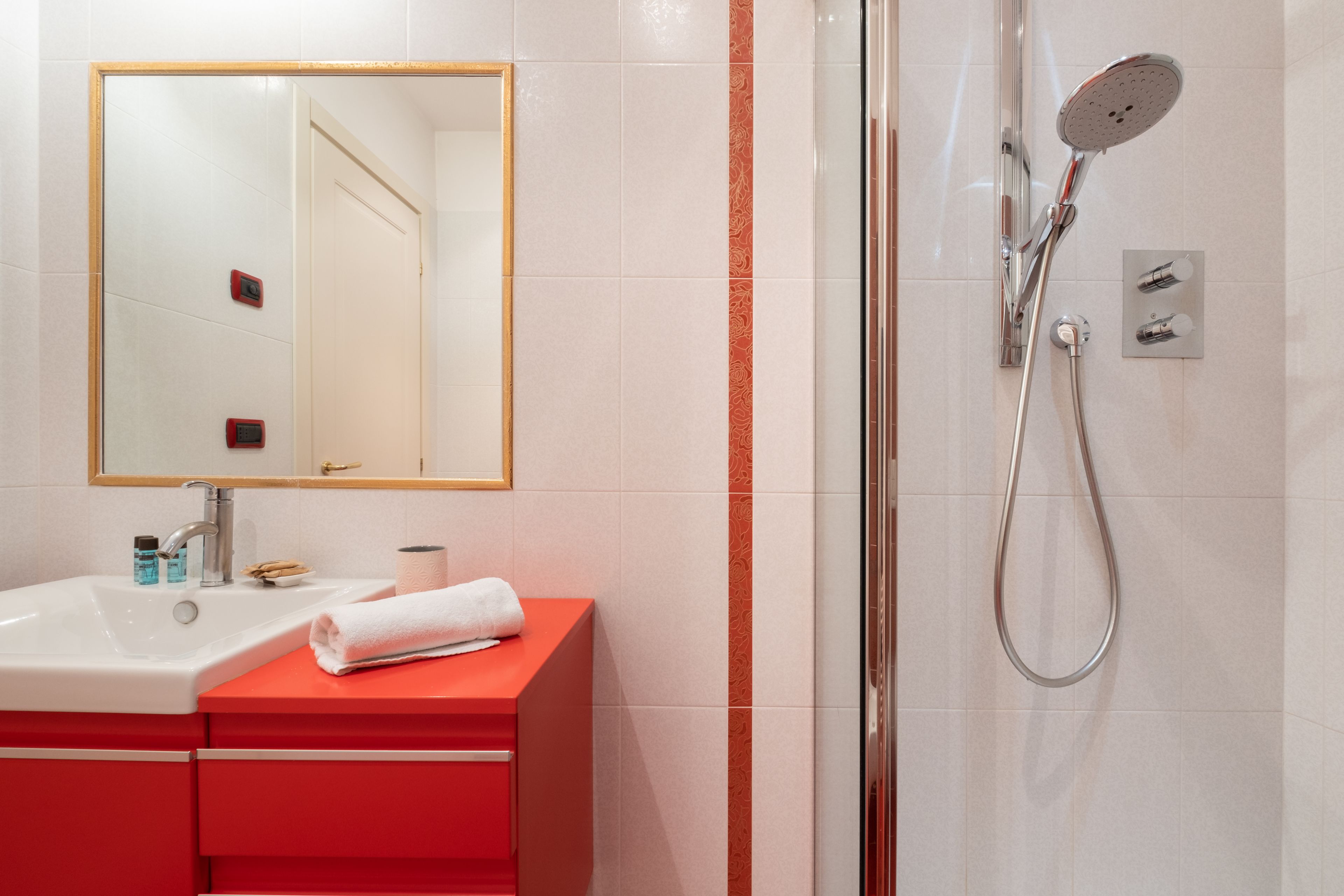 A bathroom with a white sink, red vanity, a mirror, and a shower with a handheld showerhead.