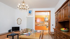 A dining area with a wooden table, chairs, and a chandelier. There is a kitchen visible in the background with orange cabinets and a window.