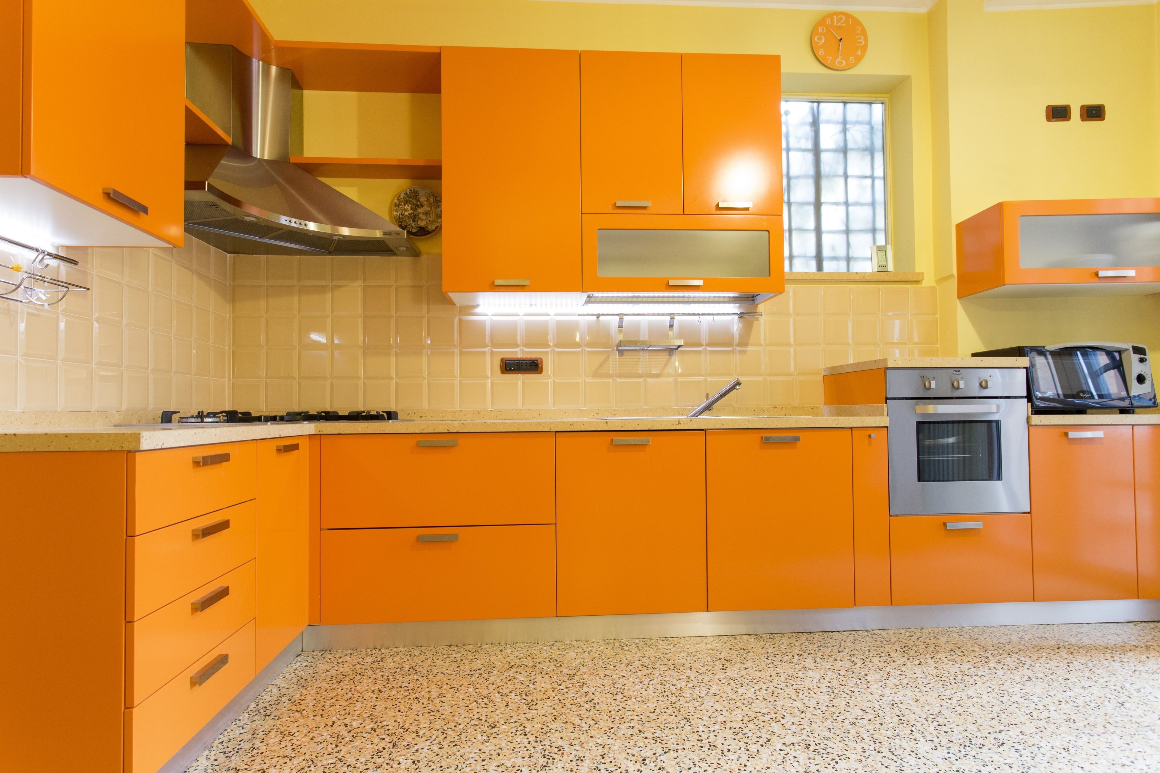 Une cuisine avec des meubles de couleur orange, une hotte de cuisinière en acier inoxydable, une horloge et une fenêtre.