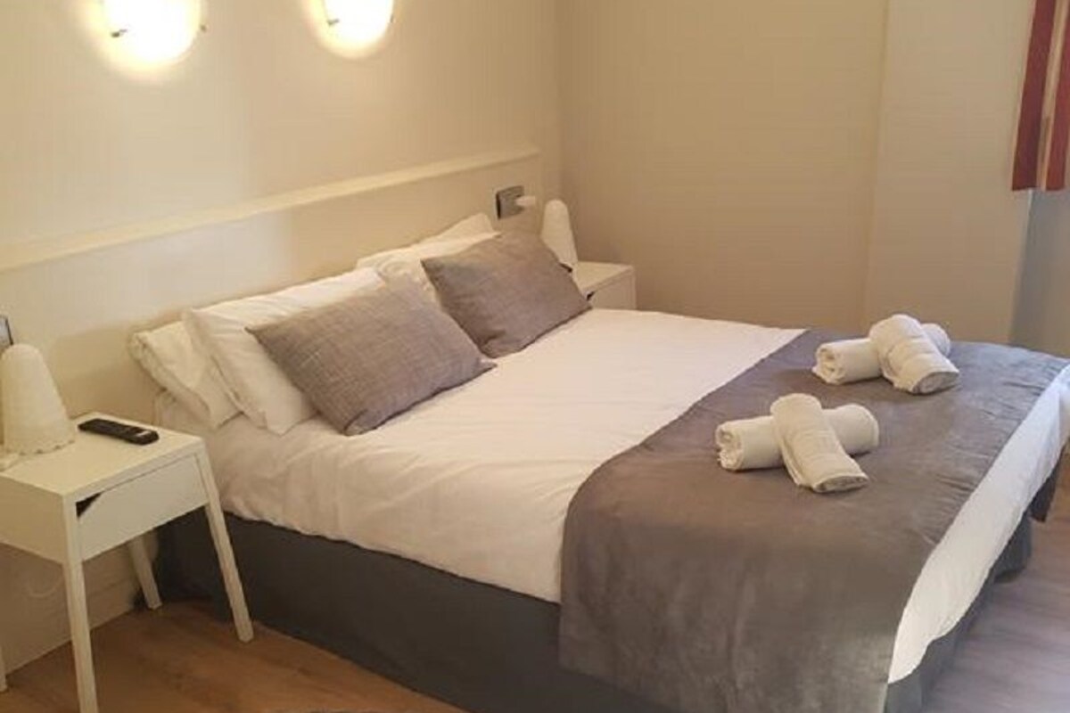 Slaapkamer met een bed, nachtkastje, twee wandlampen, een rond vloerkleed en een raam met gordijnen.