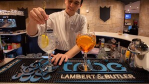 Un barman travaille dans un bar appelé Lambert & Butler. Le comptoir est bien équipé et comprend des verres, des bouteilles et un écran numérique en arrière-plan.