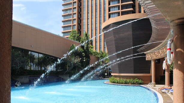 A swimming pool with water jets, surrounded by a building with balconies and a palm tree.