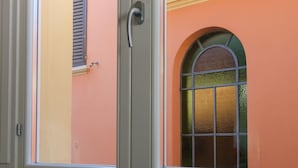 Visão pela janela com uma porta, mostrando um edifício com fachada rosa, telhado em telhas e uma antena visível.