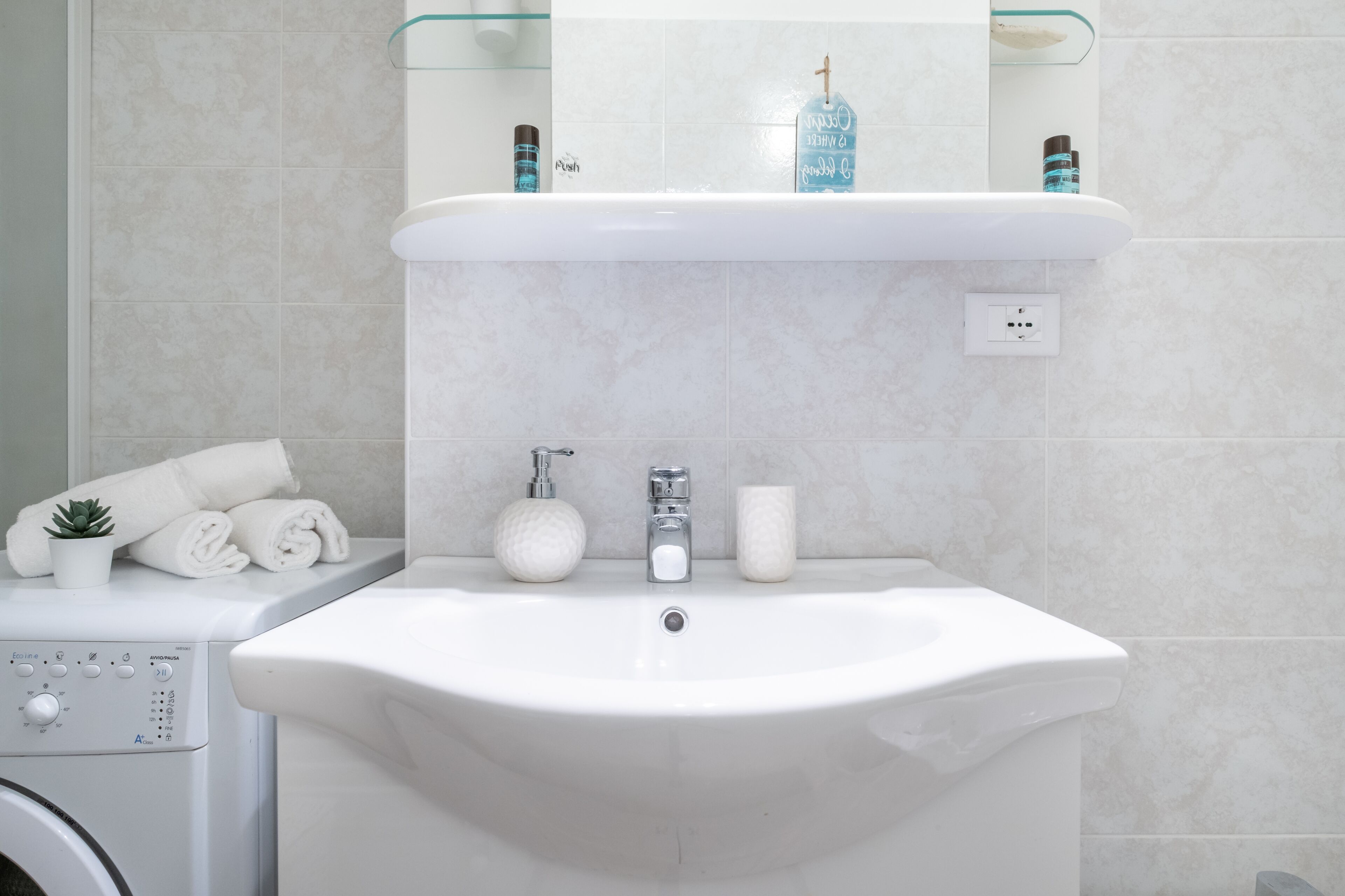A bathroom with a white sink, a glass shelf above it, and a washing machine to the left.