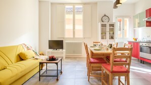 Uma sala de estar luminosa com um sofá amarelo, uma mesa de jantar com cadeiras, uma televisão e uma área de cozinha com armários e eletrodomésticos.