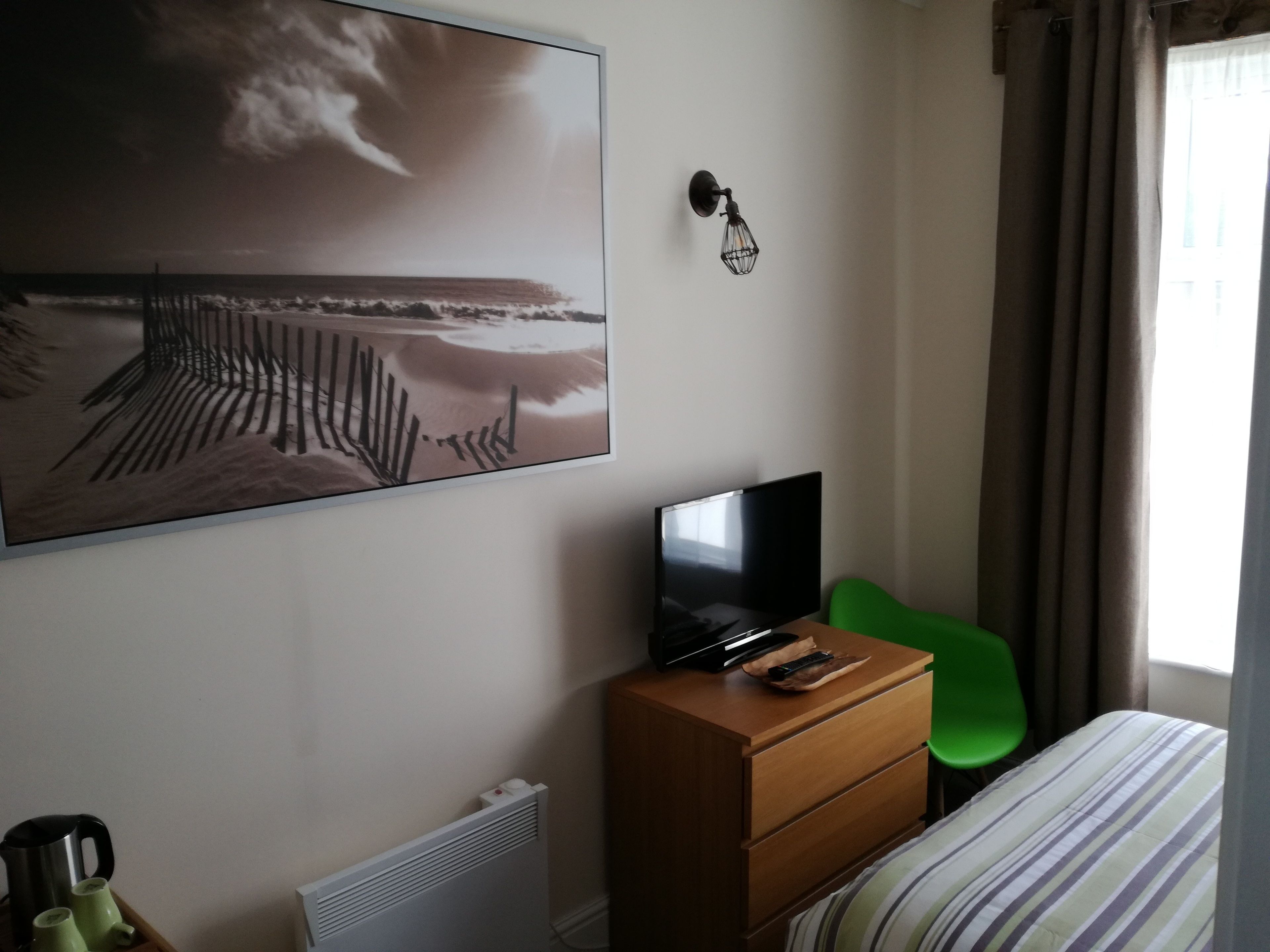 Une chambre d’hôtel avec un lit, un téléviseur posé sur une commode en bois, un fauteuil vert, un radiateur et un tableau représentant une scène de plage.