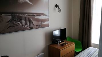 Une chambre d’hôtel avec un lit, un téléviseur posé sur une commode en bois, un fauteuil vert, un radiateur et un tableau représentant une scène de plage.