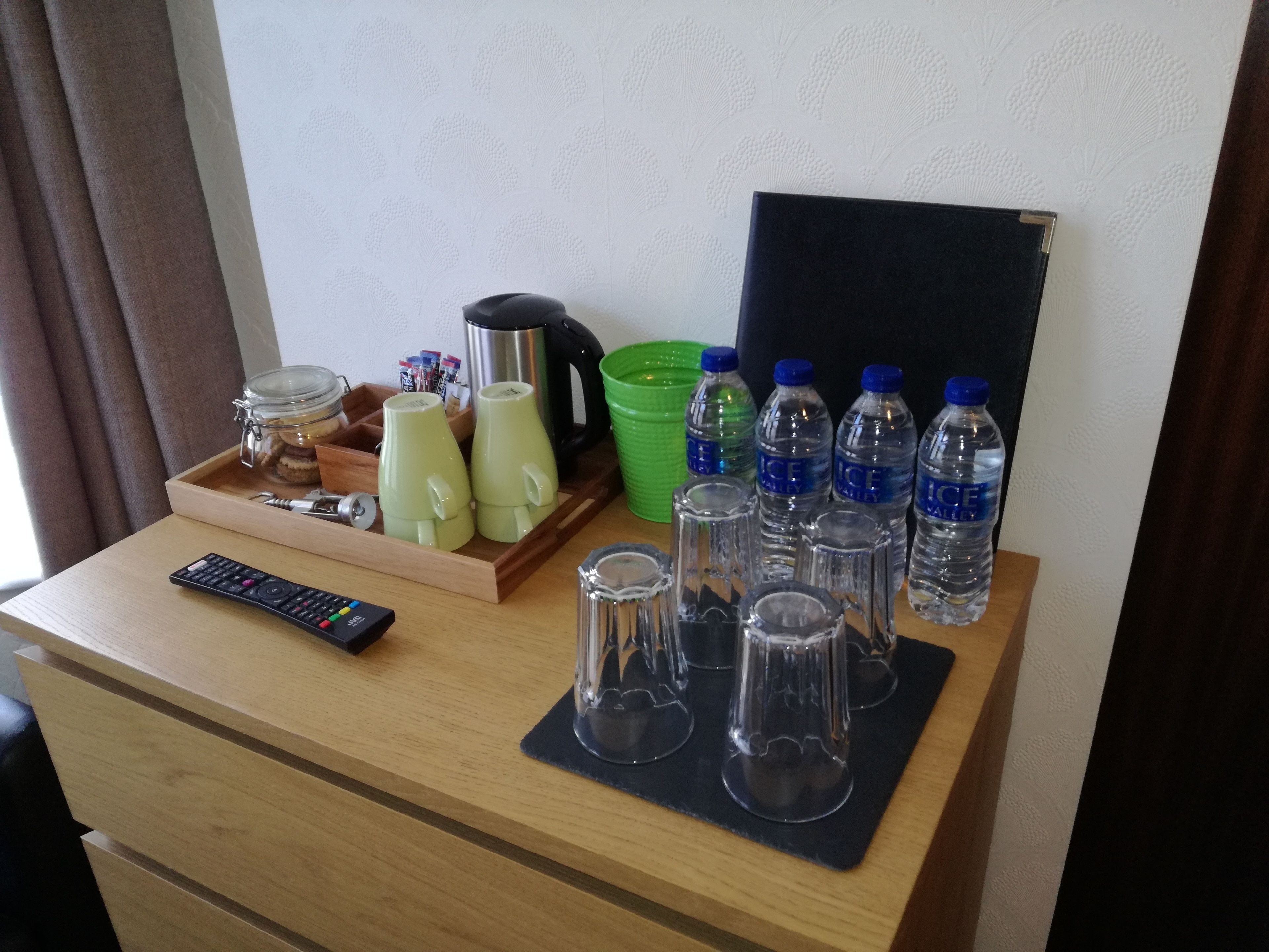 Un escritorio de madera con un control remoto, una bandeja con frascos, tazas, una tetera, un bol verde y varias botellas de agua.