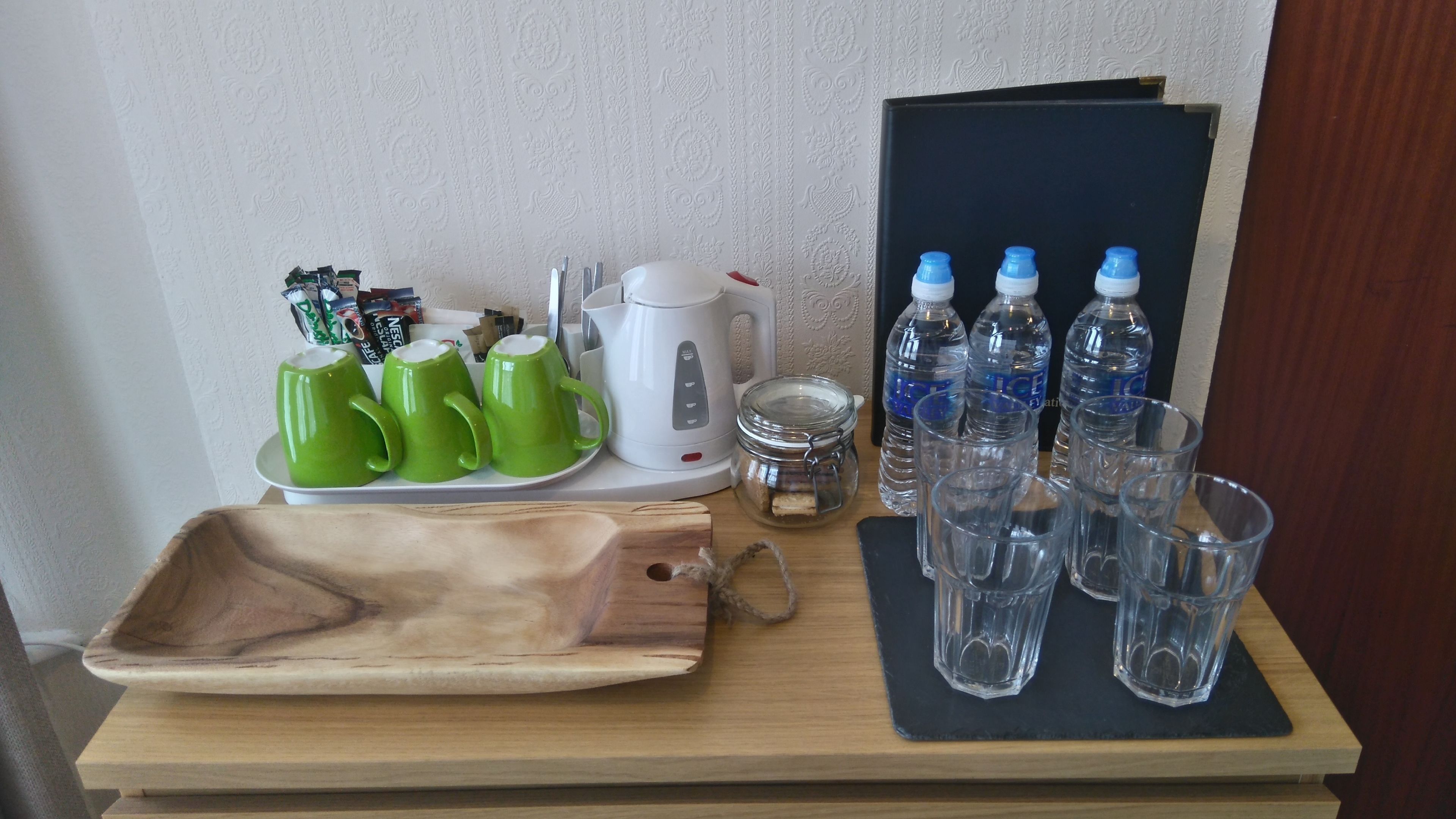 Una bandeja con tazas verdes, una cafetera blanca, un tarro de galletas y varias botellas de agua y vasos sobre una mesa de madera.