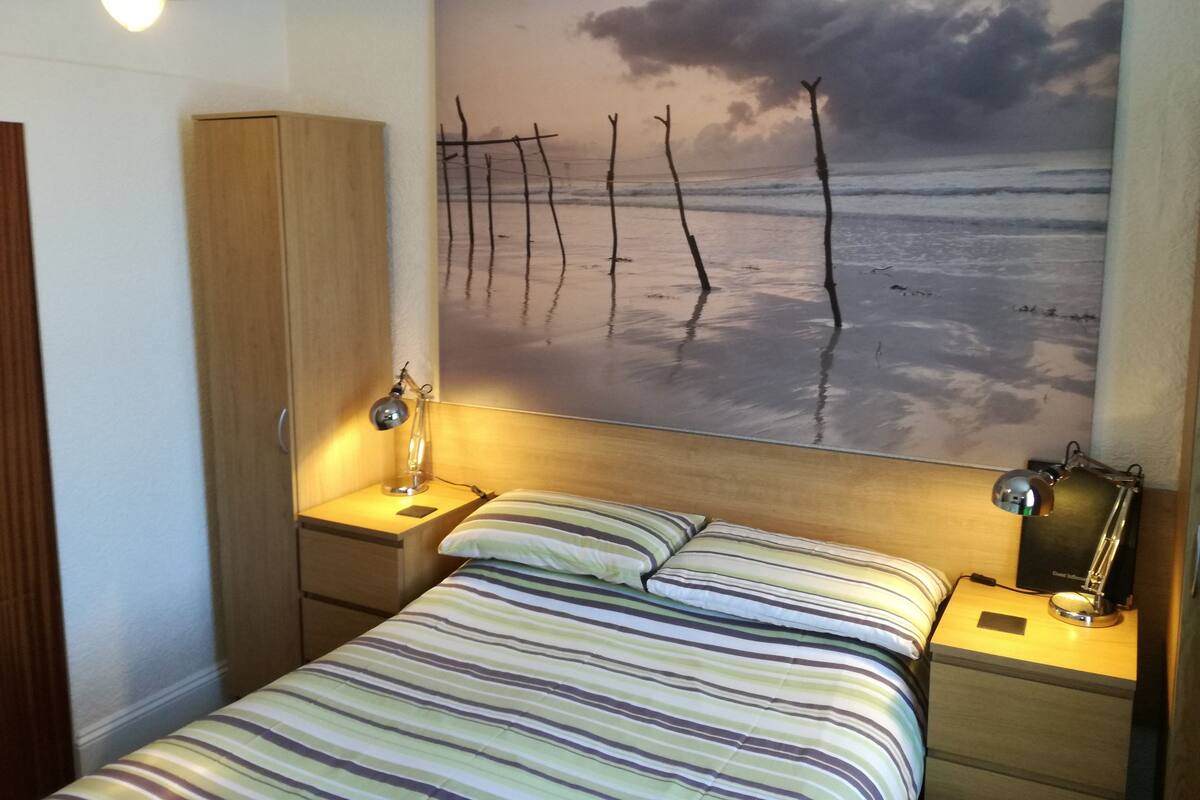 Quarto com cama, mesas de cabeceira, abajur, guarda-roupa e uma grande obra de arte na parede retratando uma praia.