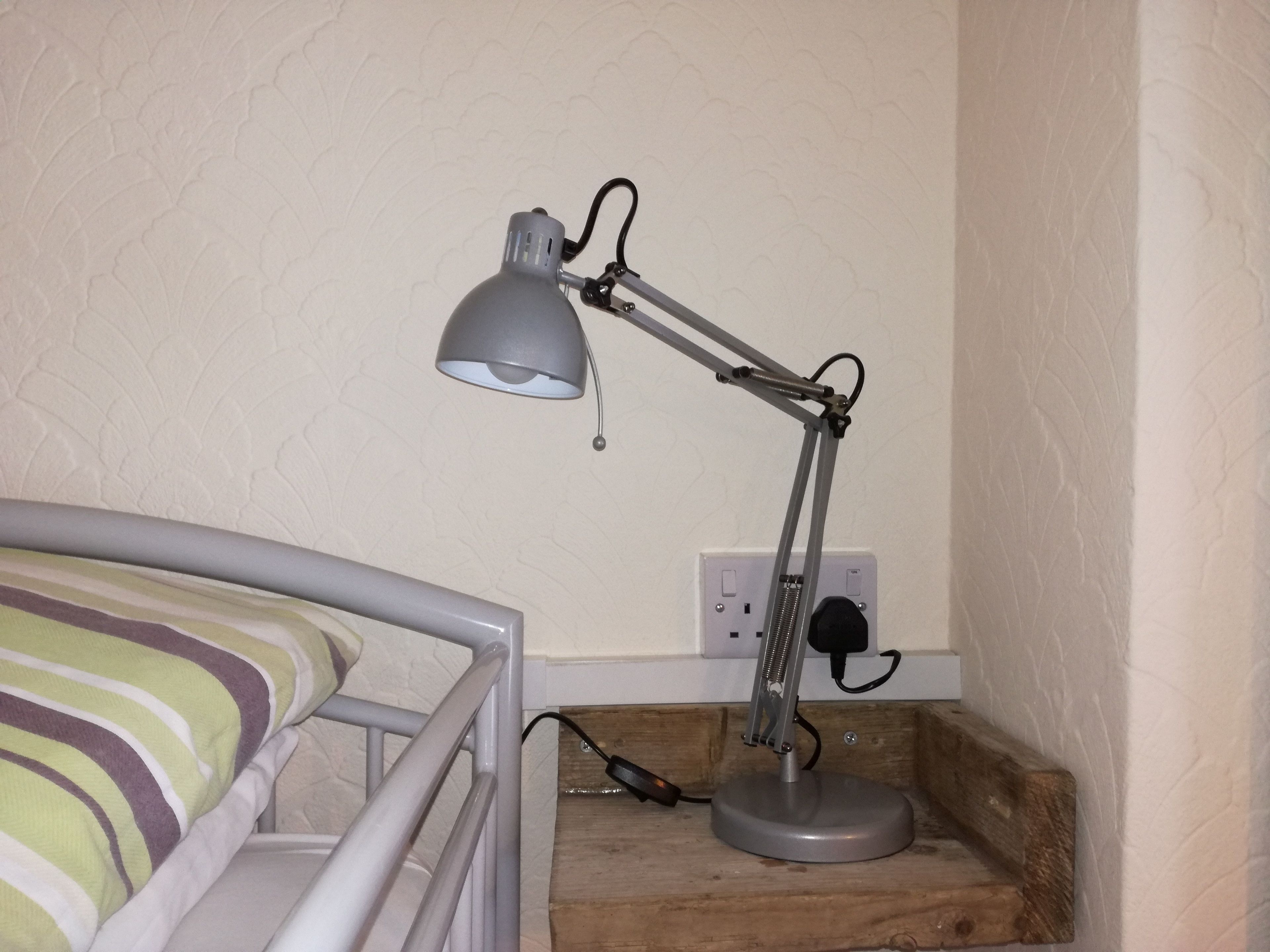 A bed with a striped pillow, a desk lamp, and a power outlet on a wooden shelf.