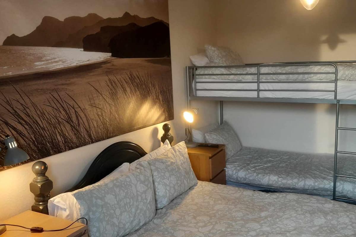 Quarto com beliche, com uma cama embaixo e outra em cima. Há uma mesinha de cabeceira com abajur e uma pequena mesa também com abajur. Um quadro de paisagem está na parede.