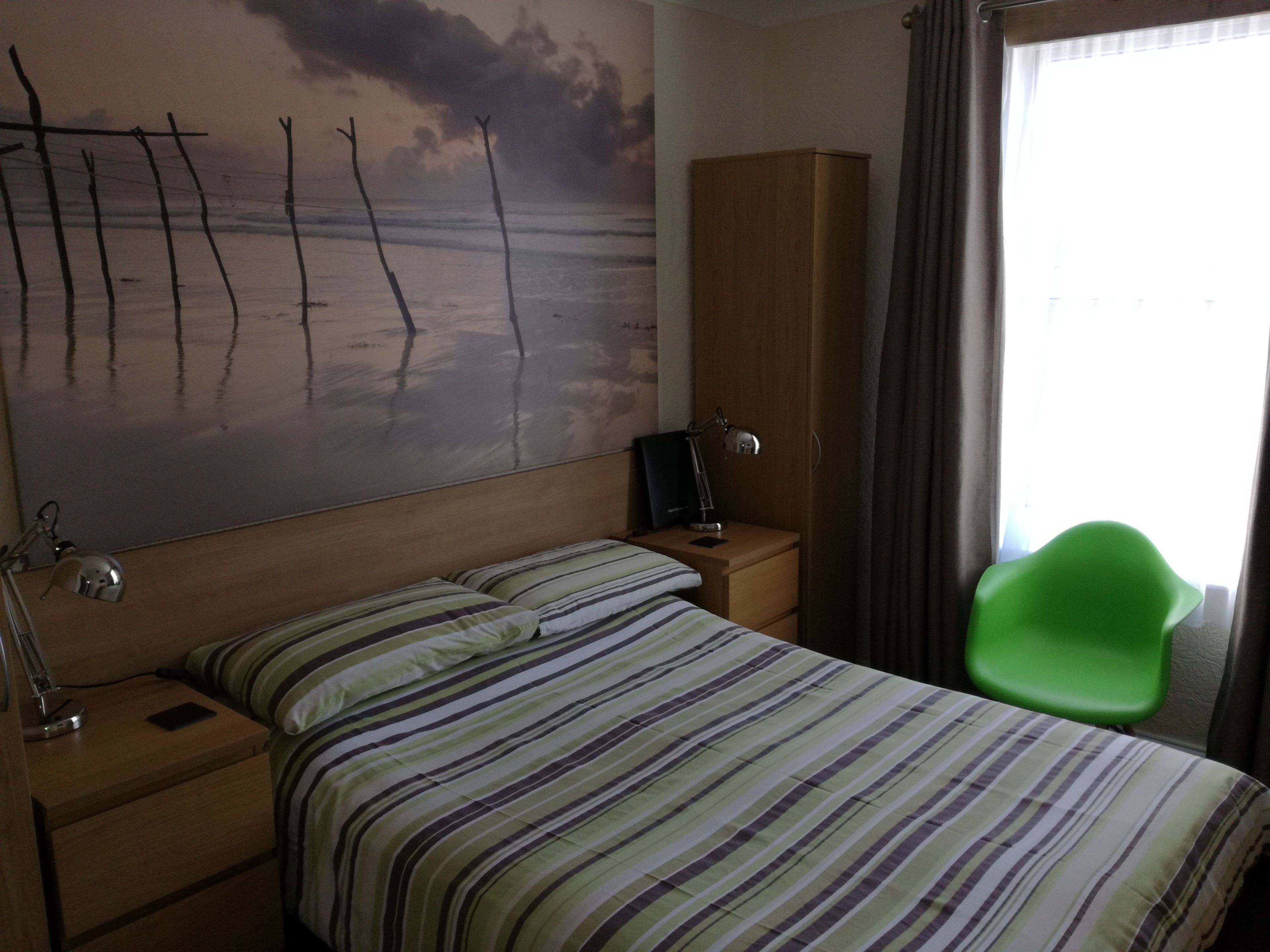 A hotel room with a bed, a nightstand, a lamp, a green chair, a wardrobe, and a painting of a beach scene.