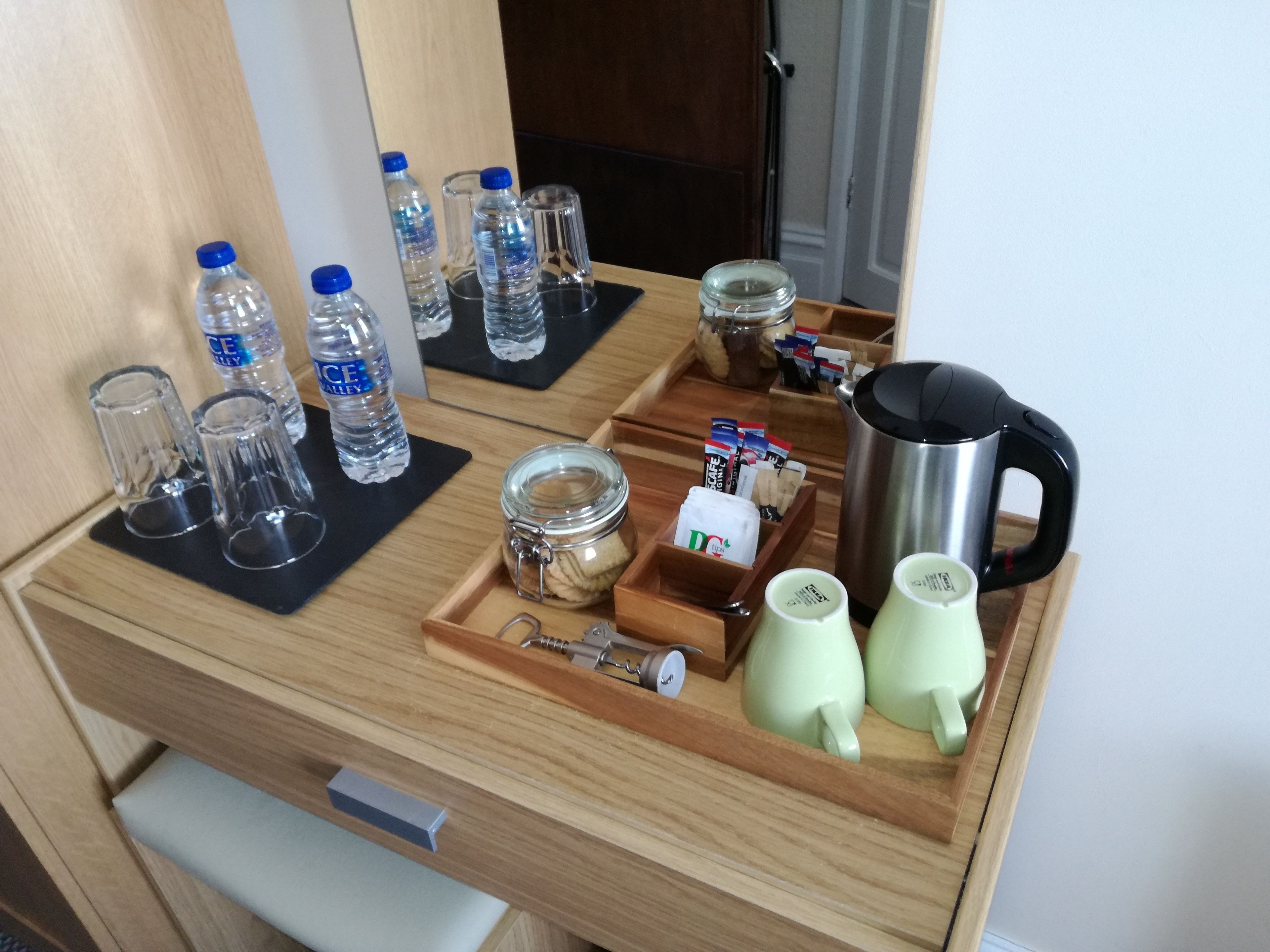 Un mini-bar d’hôtel contenant des bouteilles d’eau, des verres, un bocal de biscuits, une machine à café et des tasses à thé.