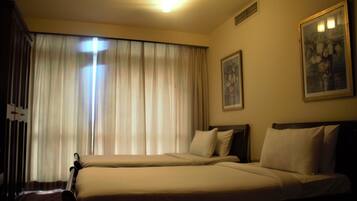 A hotel room with two beds, a large window with curtains, a ceiling light, and two framed pictures on the walls.