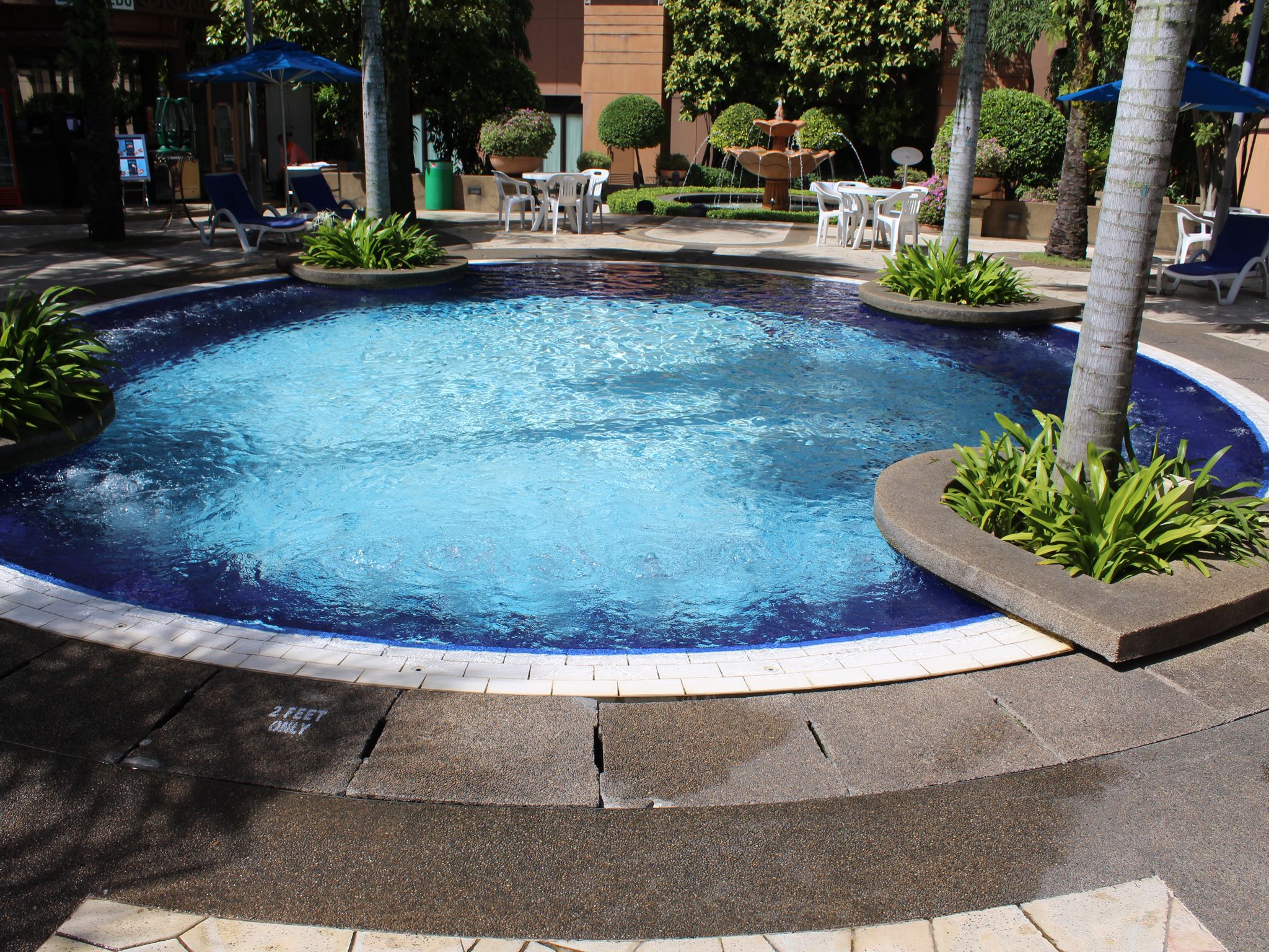A swimming pool with a fountain, surrounded by lounge chairs and tables, with a building in the background.